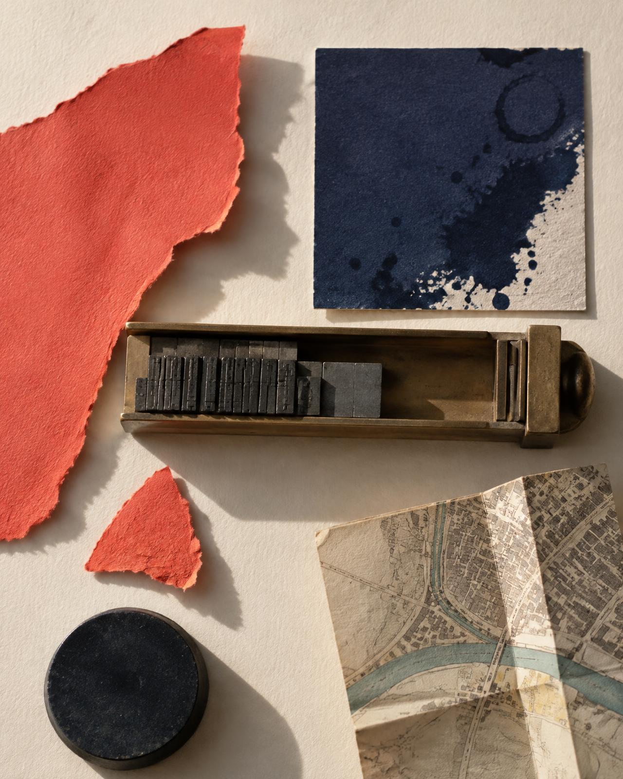 Editorial still life of torn coral paper, an ink-stained navy card, metal printing type and a folded town map